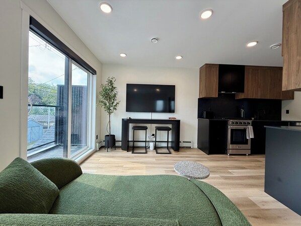 A modern living space with a large window, a flat-screen TV, a kitchen area, and a green sofa.