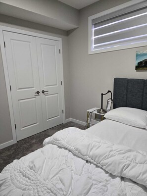 Ein Schlafzimmer mit Bett, Nachttisch mit Lampe, einem Fenster mit Jalousien und einer Tür.