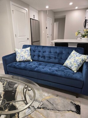 Ein Wohnzimmer mit einem blauen Sofa, zwei floralen Kissen, einem Glastisch und einem gemusterten Teppich.