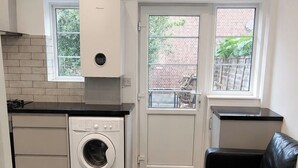 A compact kitchen with a washing machine, a black leather sofa, and a trash bin. - Serviced Townhome: 4 bedroom, 3 bathroom+ garden (London)