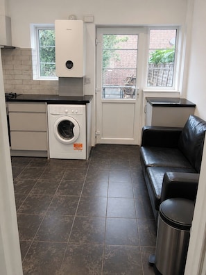 A compact kitchen with a washing machine, a black leather sofa, and a trash bin. - Serviced Townhome: 4 bedroom, 3 bathroom+ garden (London)