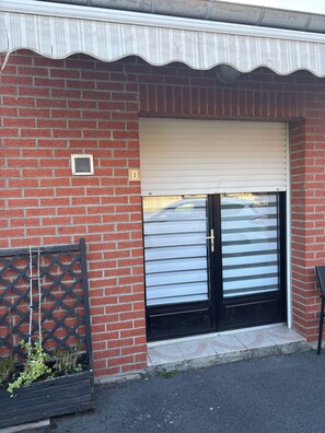Ein Backsteinhaus mit heruntergelassenem Rolltor, einem kleinen Fenster mit Metallgitter und einem schwarzen Metallzaun mit Bepflanzung.