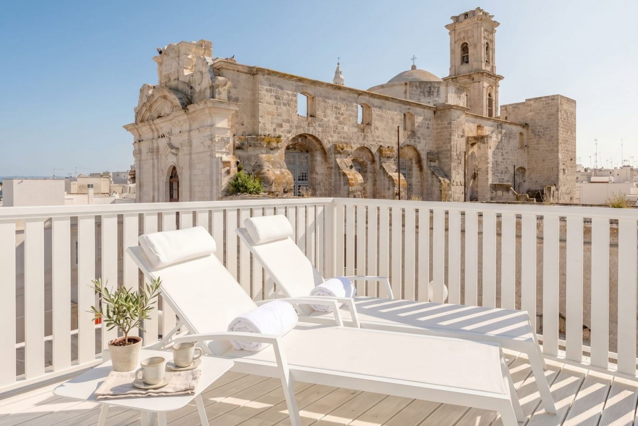 A rooftop terrace with white lounge chairs and a table, overlooking a historic stone building with arches and a bell tower.