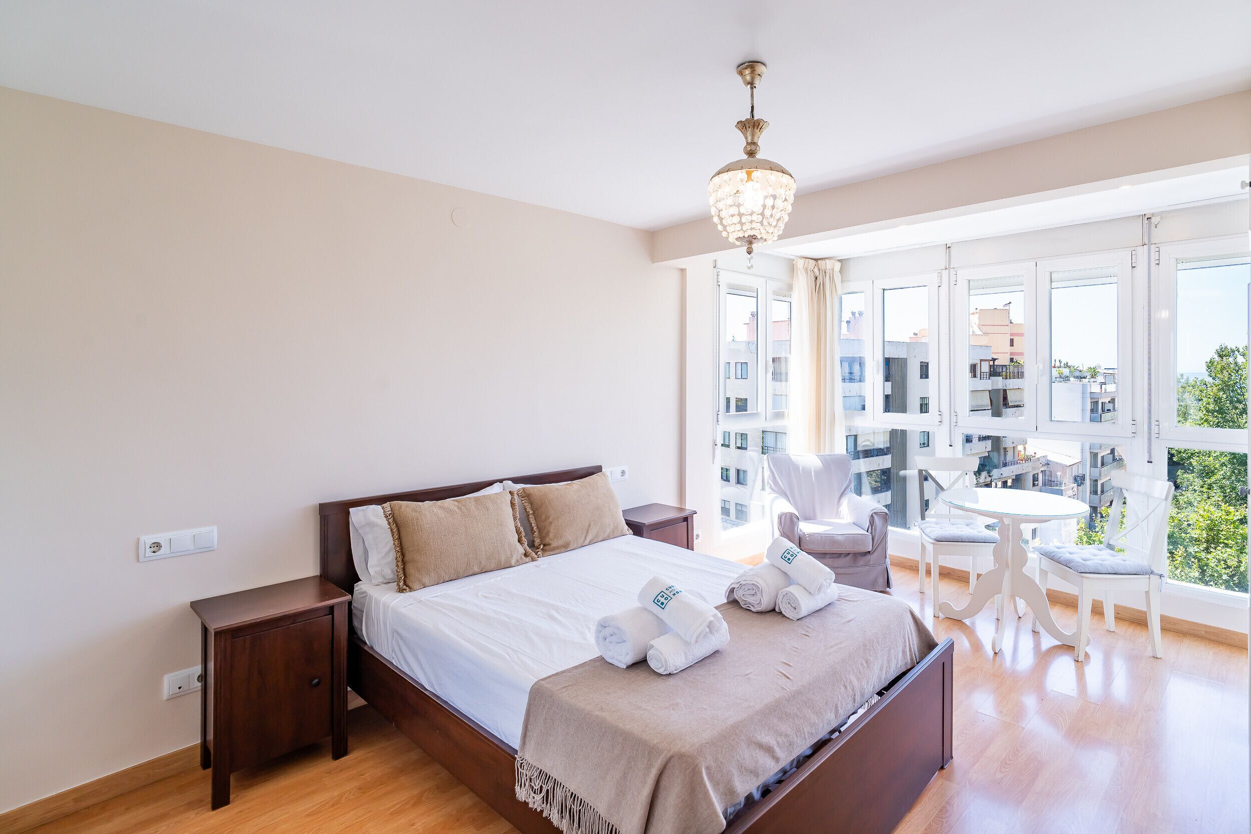 Une chambre d’hôtel comprenant un lit, des tables de chevet, un lustre, un balcon avec vue sur les bâtiments et une table avec des chaises.