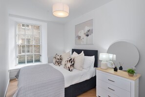 A bedroom with a large window, a bed with patterned pillows, a bedside table with a lamp, and a wall-mounted mirror. - Modern Apartment in City Centre (Edinburgh)