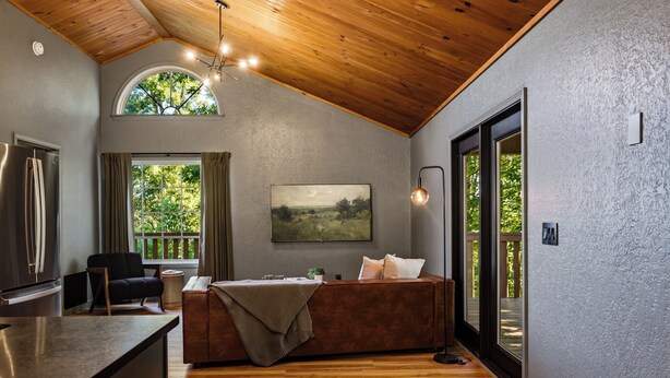 A modern interior with a wooden ceiling, a kitchen area, a seating area with a sofa and a table, and a large window providing a view of greenery. - NEW Couples! VIEW, Barrel Sauna, Hot Tub, Fire Pit (Sevierville)