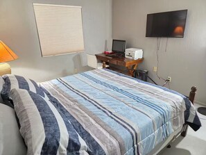 A bedroom with a bed, a desk with a computer, a television on the wall, and a window with blinds. - Peaceful 2-bedroom condo with AC in sunny Tucson (Tucson)