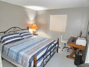 Quarto com cama, escrivaninha, cadeira, abajur e janela.