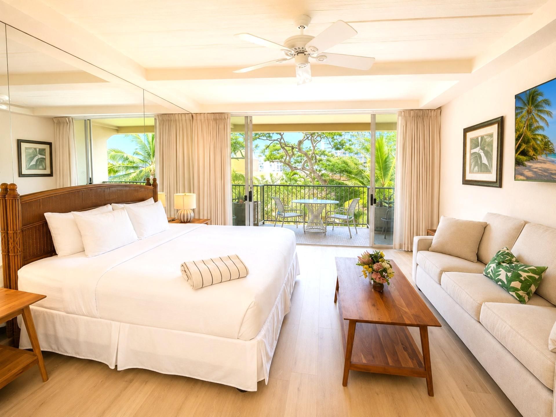 A hotel room with a large bed, a sofa, a coffee table, and a balcony with a view of palm trees.