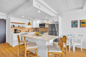A modern kitchen with white cabinetry, a central island, wooden chairs, and a dining table. - Whaler | 3 Oceanfront Villas, Sleeps 6 | Car Incl. w/ 6+ Nights | ML-4229 by KBM (Lahaina)