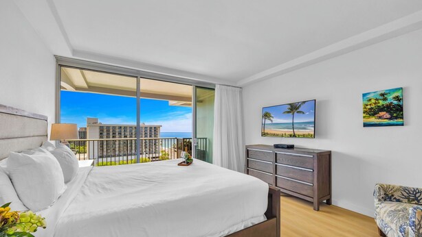Habitación de hotel con una cama grande, televisión, cómoda y balcón con vistas a edificios y al mar.