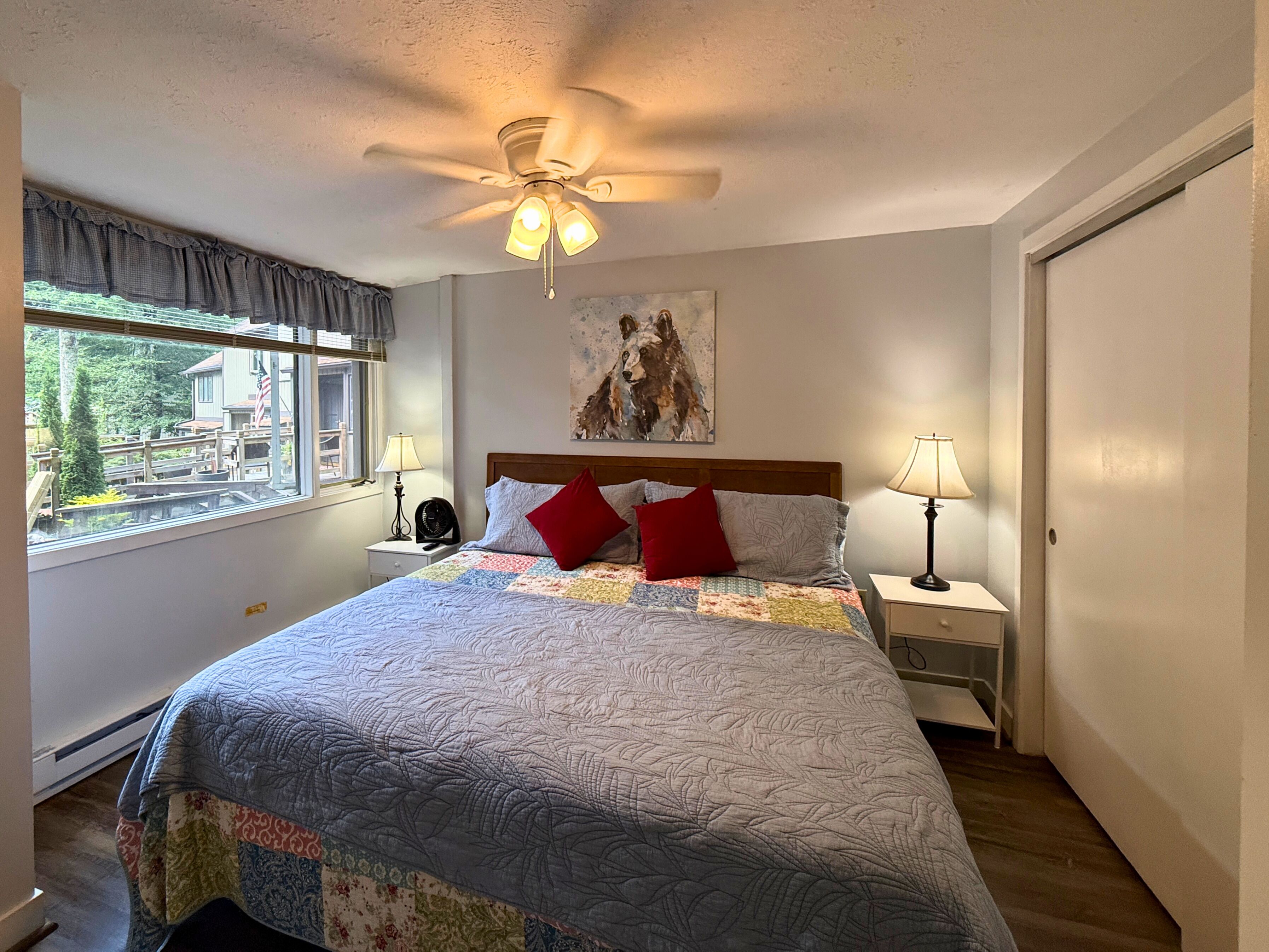 Ein Schlafzimmer mit einem Bett, zwei Nachttischen mit Lampen, einem Deckenventilator, einem Fenster mit Vorhängen und einem Gemälde an der Wand.