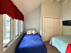 Ein Schlafzimmer mit einem Einzelbett, einem Fenster mit Vorhängen, einer Tür und einem an der Wand befestigten Fernseher.
