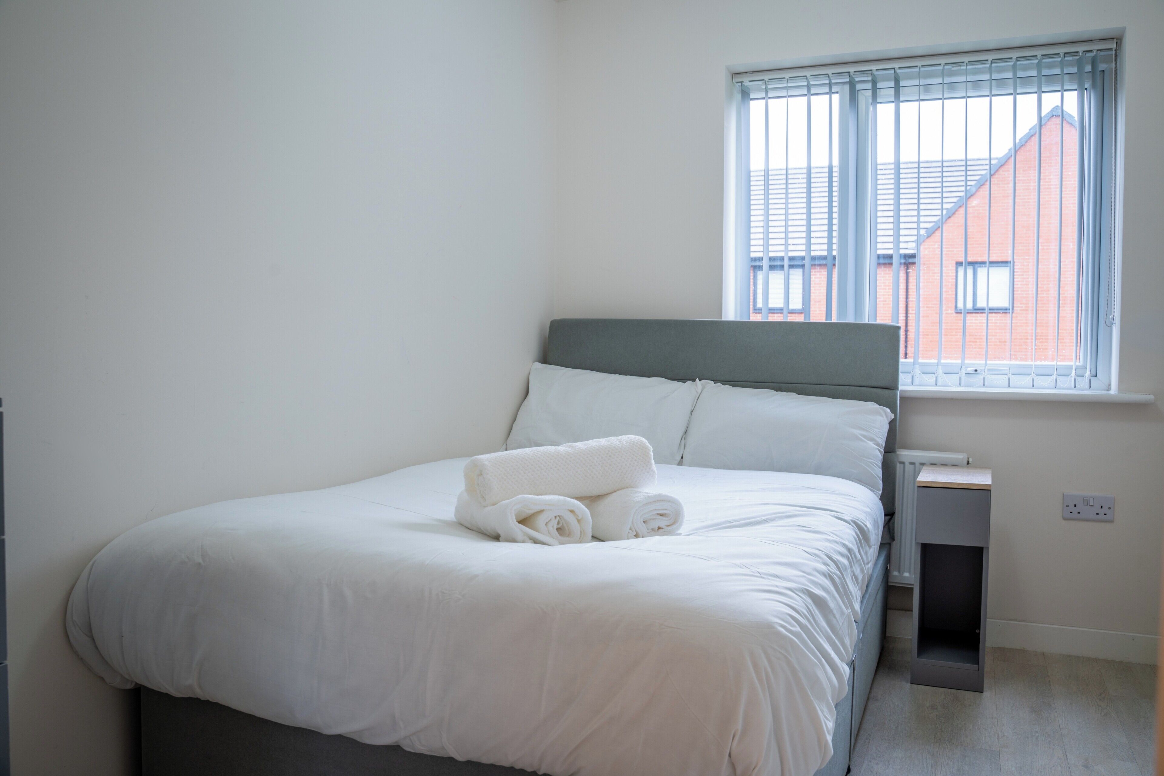 Een netjes opgemaakt bed met witte lakens en opgerolde handdoeken, een grijs hoofdbord en een raam met jaloezieën dat uitkijkt op een gebouw.