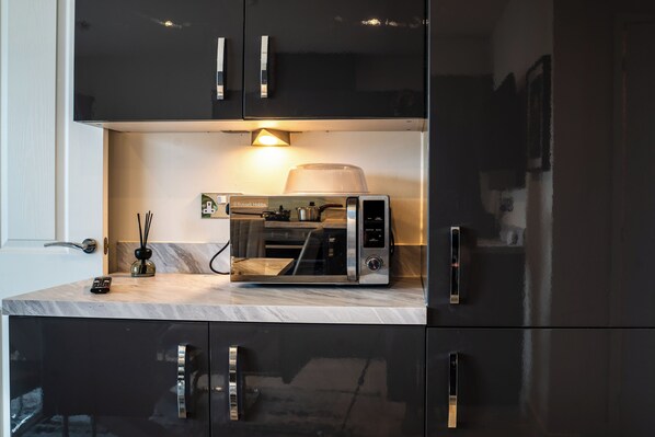 Una cocina con horno microondas integrado, cafetera y una pequeña mesa con un control remoto y un objeto decorativo.