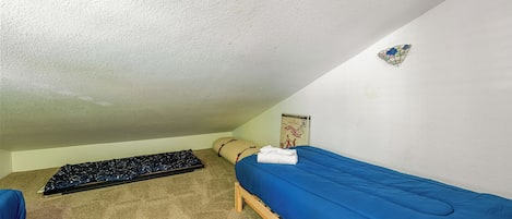 Une petite chambre avec un lit simple, un matelas bleu, un cadre en bois, une moquette beige et une petite chaise bleue.