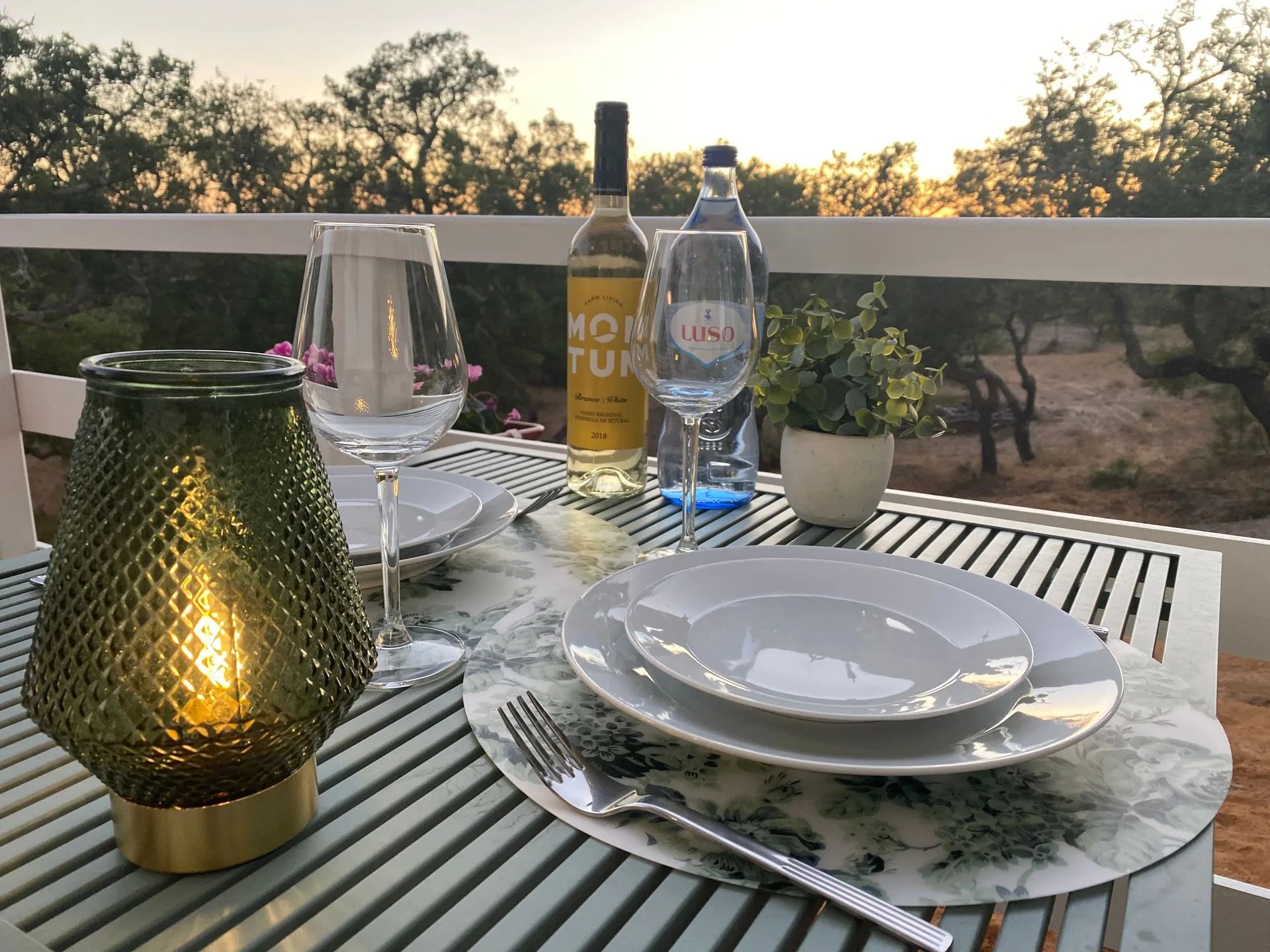 Ein Tisch ist gedeckt mit Tellern, Besteck, einer Weinflasche, Gläsern und einer dekorativen Lampe auf einer Terrasse im Freien mit Blick auf Bäume.