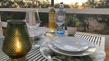 Un tavolo apparecchiato con piatti, posate, una bottiglia di vino, bicchieri e una lampada decorativa su una terrazza esterna con vista sugli alberi.
