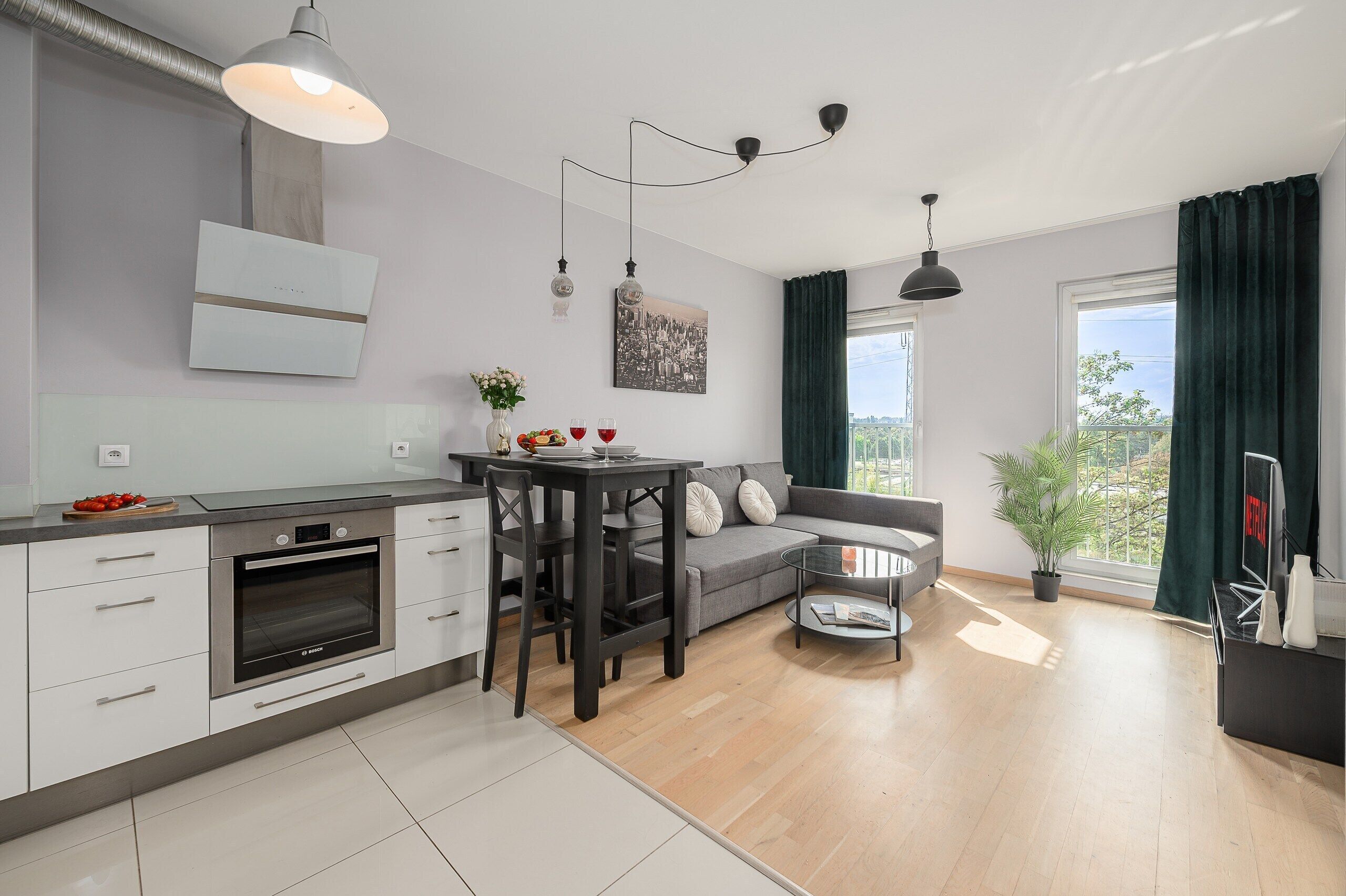 Une cuisine moderne équipée d’un four Bosch, une table à manger avec des chaises, un canapé gris et une grande fenêtre avec des rideaux.