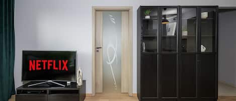 Una habitación con una puerta de madera, un mueble negro con un televisor que muestra 'NETFLIX' y otro mueble negro con puertas de cristal que contiene libros y objetos decorativos.