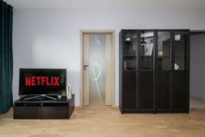 A room with a wooden door, a black cabinet with a television displaying 'NETFLIX', and a black cabinet with glass doors containing books and decorative items. - Lumina 2-room Apart with Balkony & Metro (Warszawa)