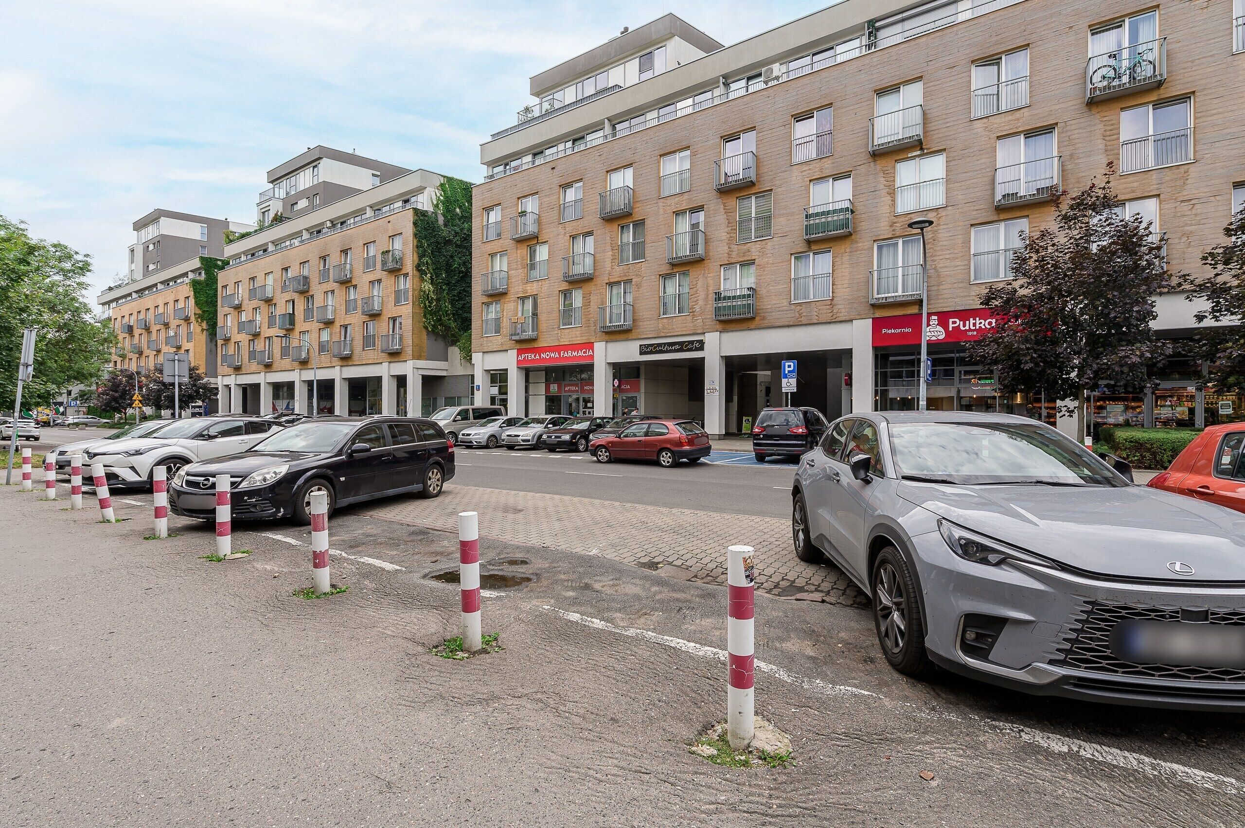 A multi-story building with a pharmacy and cafe on the ground floor, parked cars, and a pedestrian walkway.