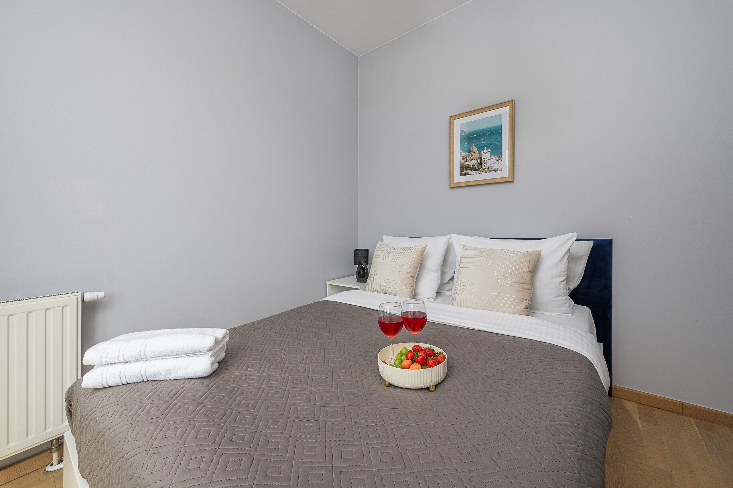 Un dormitorio con cama, almohadas blancas, una mesita de noche con lámpara, un cuadro en la pared y un frutero sobre la cama.