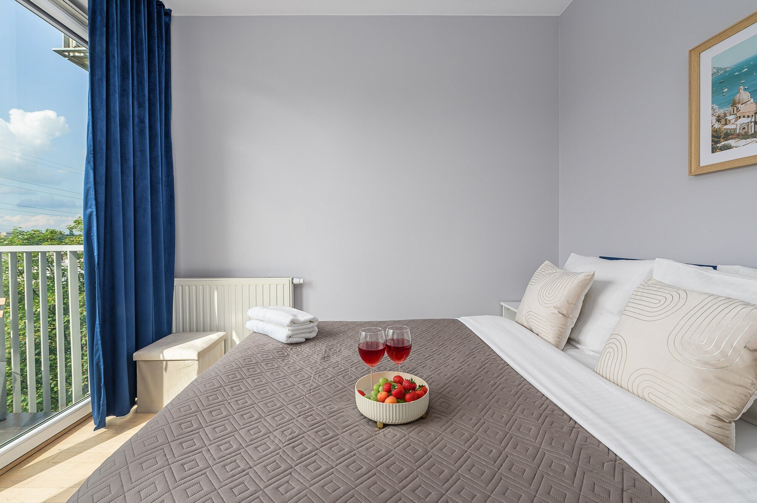 A hotel room with a bed, a bowl of fruit, two glasses of red wine, and a painting on the wall.