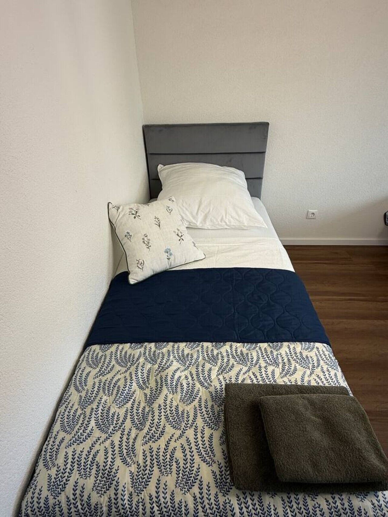 Ein Einzelbett mit einer dunkelblauen Steppdecke, einem weißen Kissen mit Blumenmuster und einem braunen Handtuch.