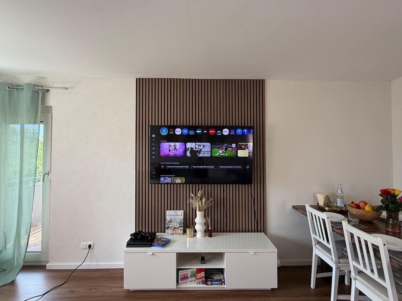 Un salon avec un téléviseur fixé au mur, un meuble de rangement blanc et un coin repas comprenant une table et des chaises.