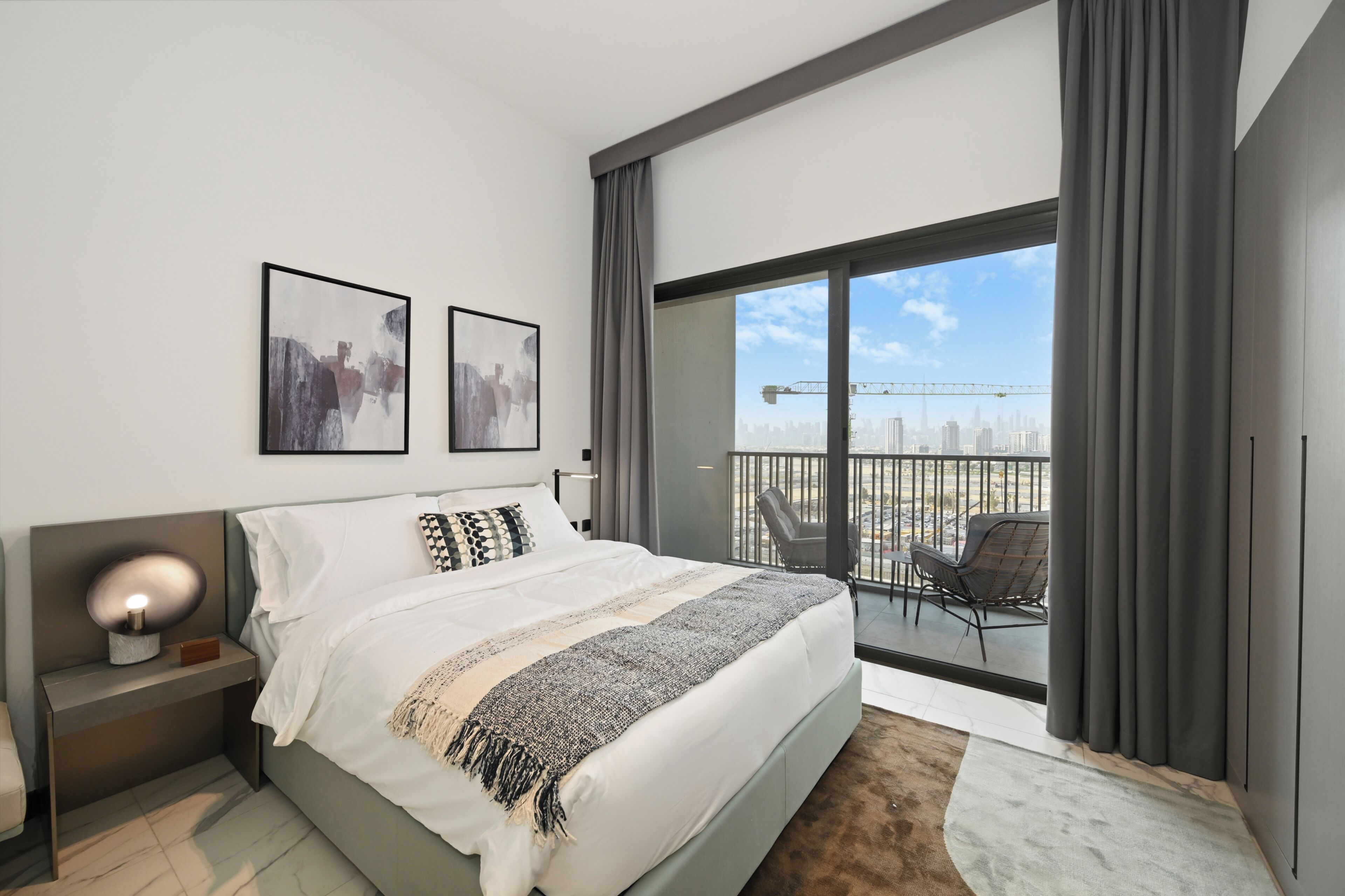 A modern hotel room with a large bed, two framed artworks, a nightstand, a balcony with a view of the city, and a city skyline.