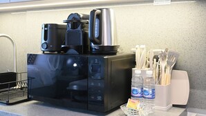Cozinha com micro-ondas, cafeteira e chaleira na bancada. Utensílios em suporte e duas garrafas de água.
