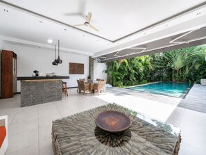 Eine moderne Küche mit einer Steinen Insel, Essbereich mit Korbstühlen, Deckenventilator und Blick auf einen Pool mit üppiger Bepflanzung.