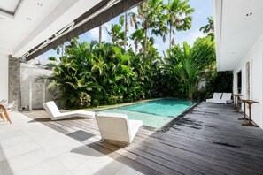 Un espace piscine extérieur moderne, doté d’une terrasse en bois, de chaises longues blanches et d’une végétation tropicale luxuriante.