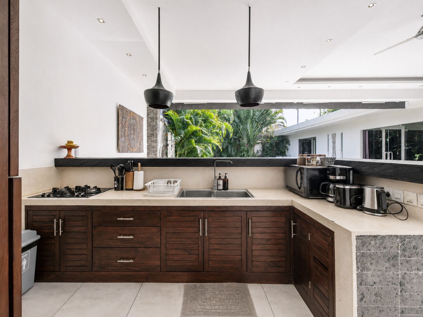 A modern kitchen with a central island, dark wooden cabinets, a stainless steel sink, and a view of greenery outside.