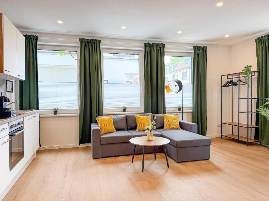 A modern living room with a grey sectional sofa, a round coffee table, and a kitchen area with white cabinets.