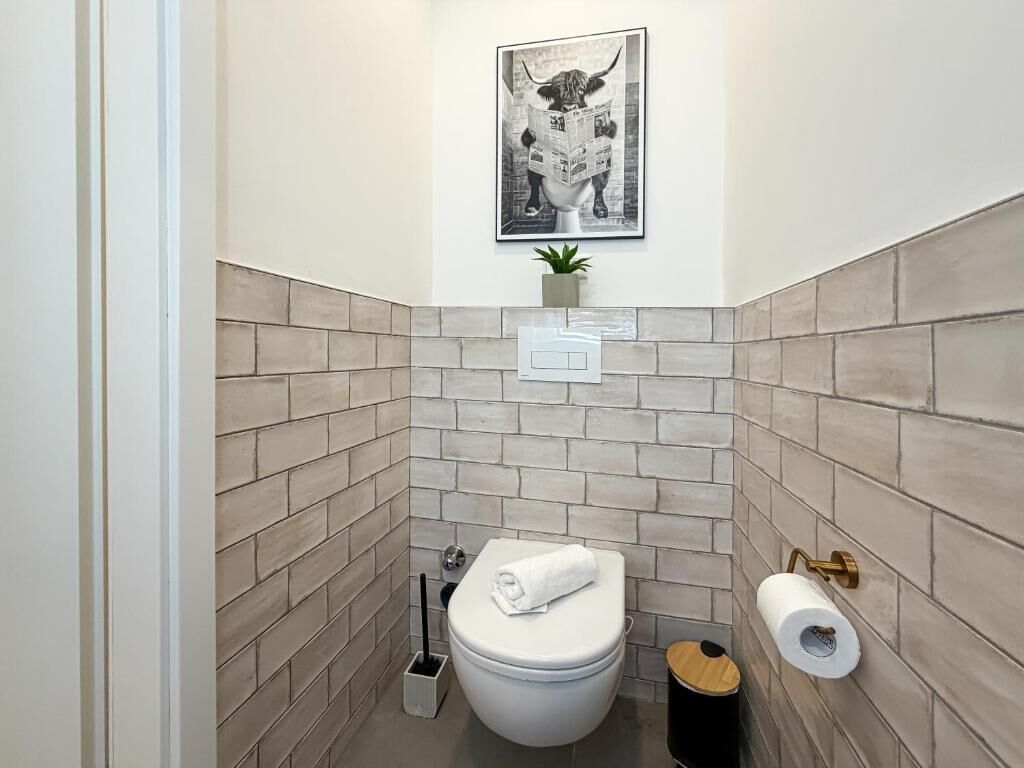 A small bathroom with a toilet, a wall-mounted toilet paper holder, a wall-mounted light, and a framed picture above the toilet.