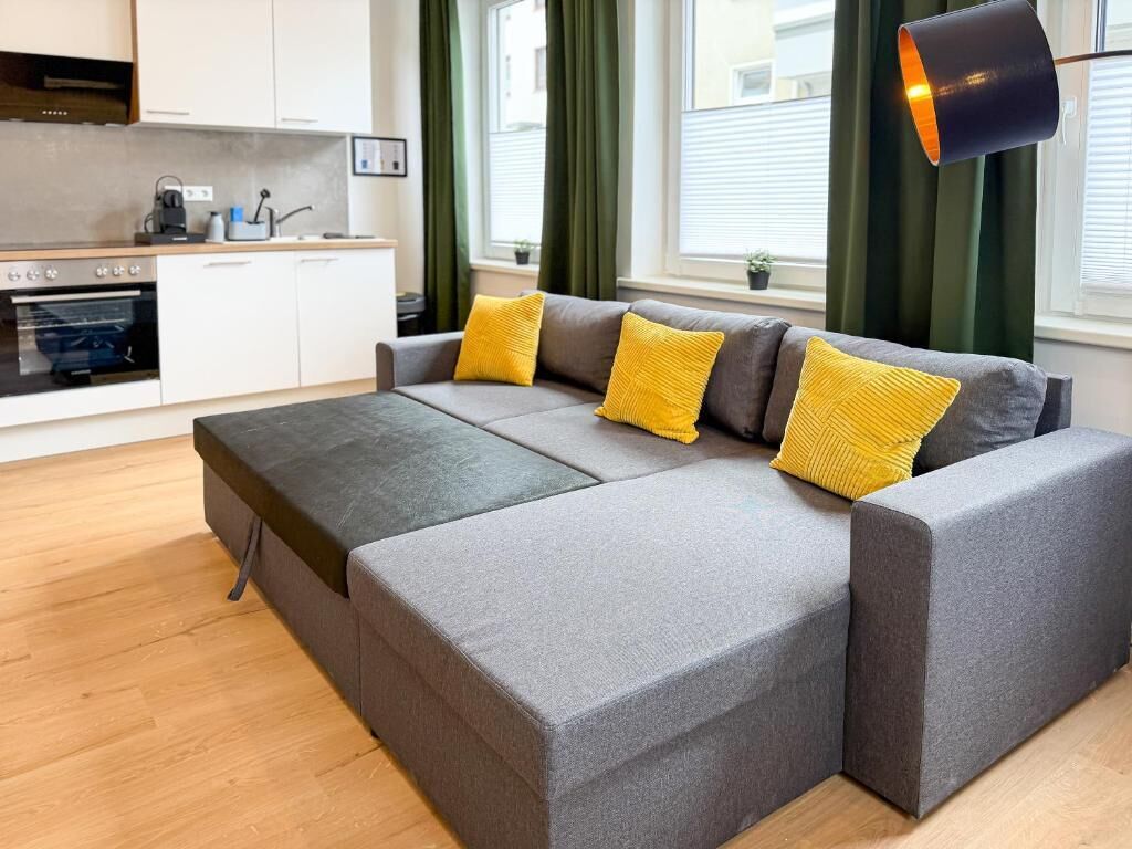 Un espacio habitable moderno con sofá, zona de cocina y ventana con cortinas.