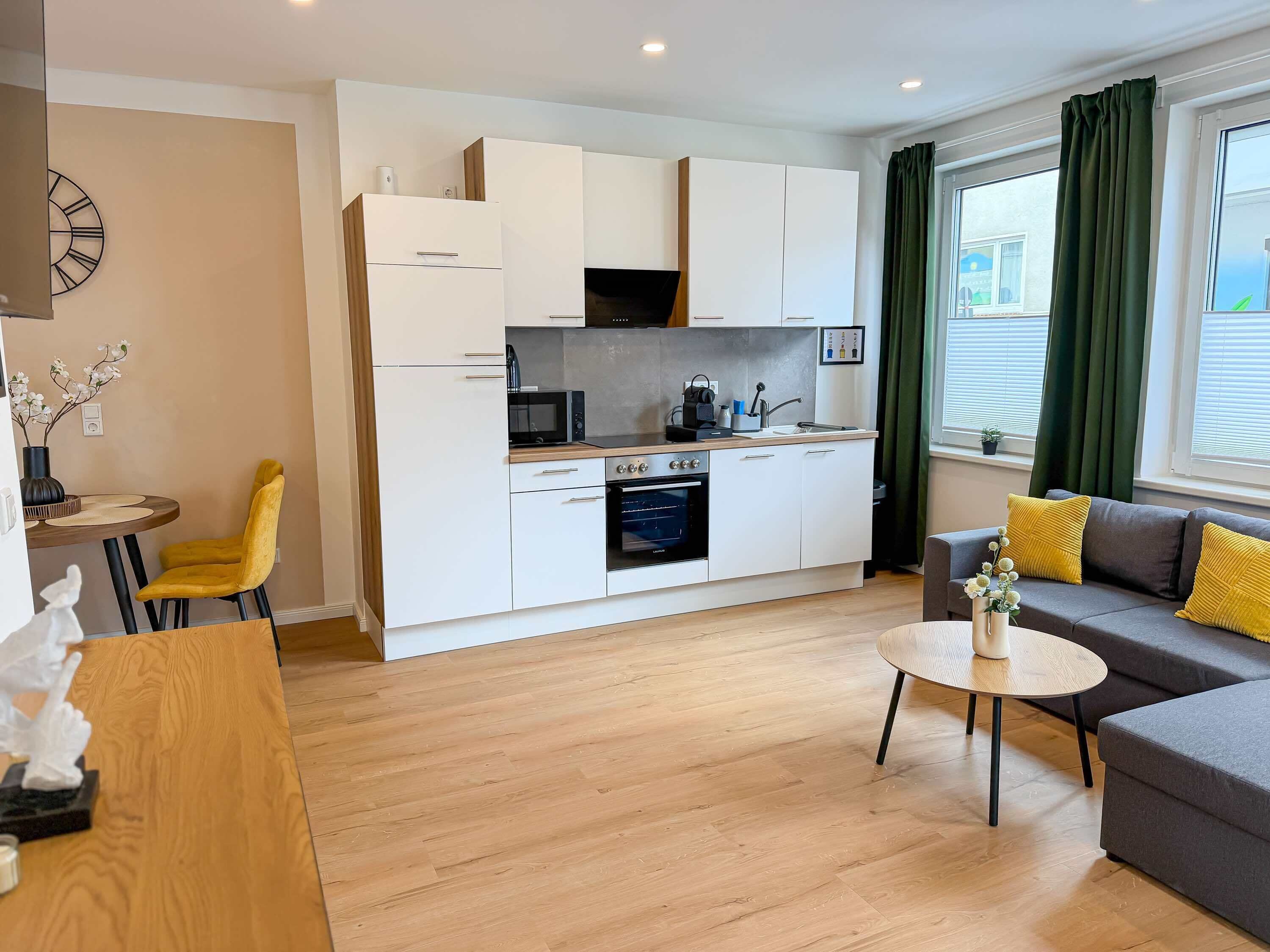 A modern kitchen with white cabinets, a microwave, and an oven. There is a dining area with a yellow chair and a round table. A gray sofa with yellow cushions is in the living area.