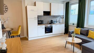 A modern kitchen with white cabinets, a microwave, and an oven. There is a dining area with a yellow chair and a round table. A gray sofa with yellow cushions is in the living area.