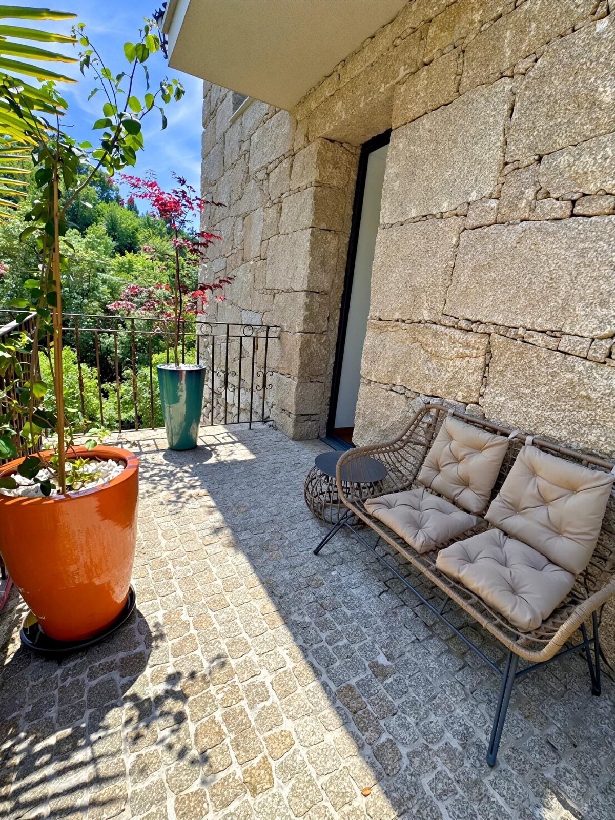 Un mur de pierres, un canapé en osier avec des coussins et une plante en pot sur une terrasse dallée.