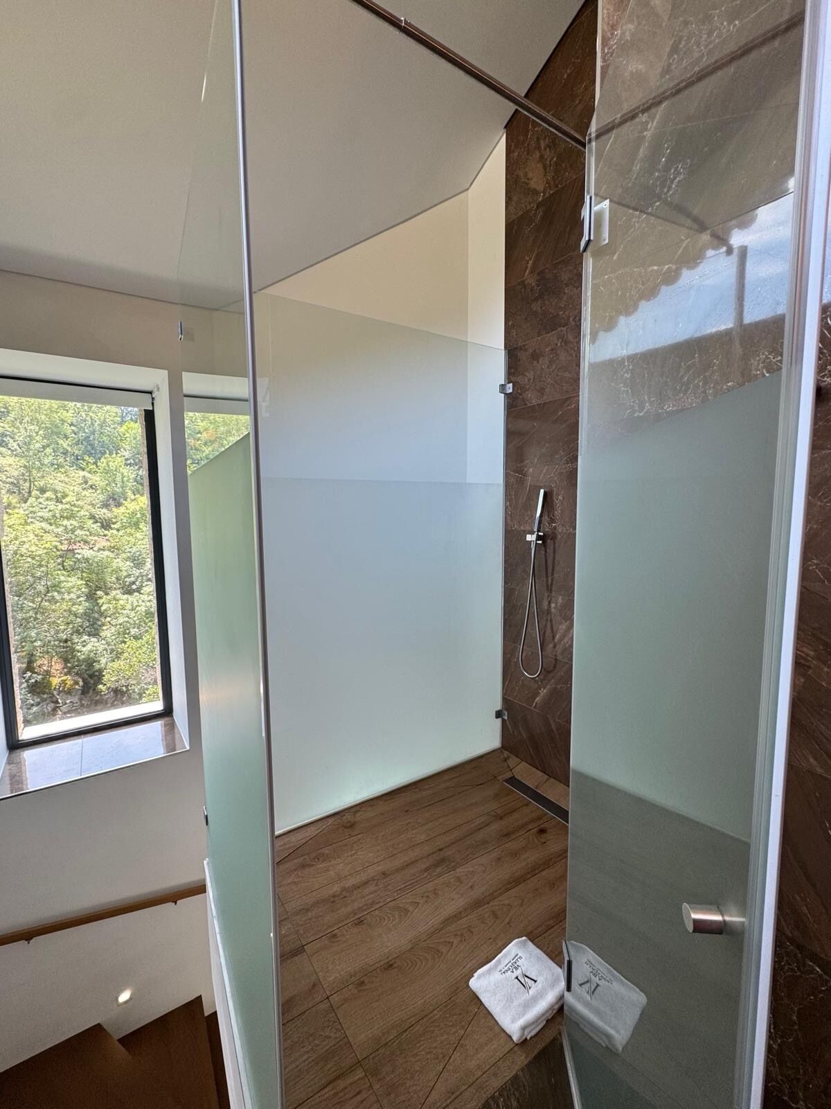 Une salle de bain moderne avec une douche à l’italienne en verre, un plancher en bois et une fenêtre donnant sur la verdure à l’extérieur.
