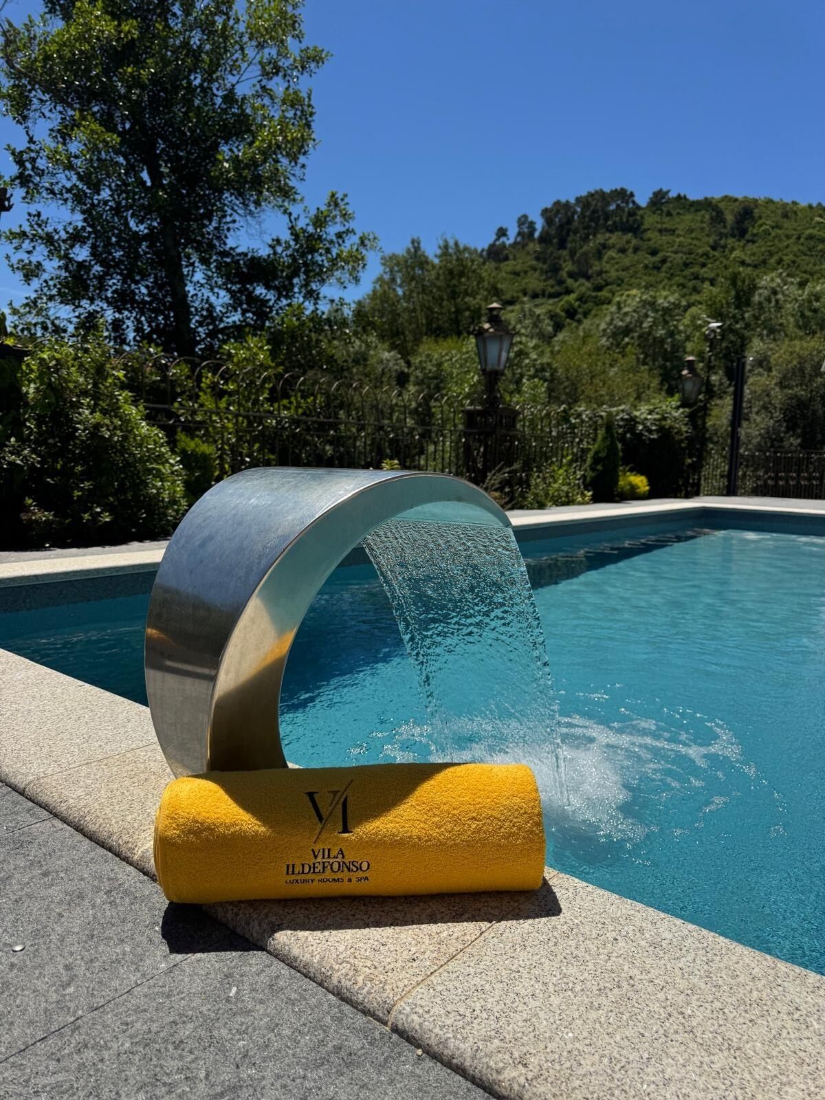 Une fontaine ou un élément décoratif aquatique doté d’une structure métallique et d’une base jaune, placée au bord d’une piscine et entourée de verdure.