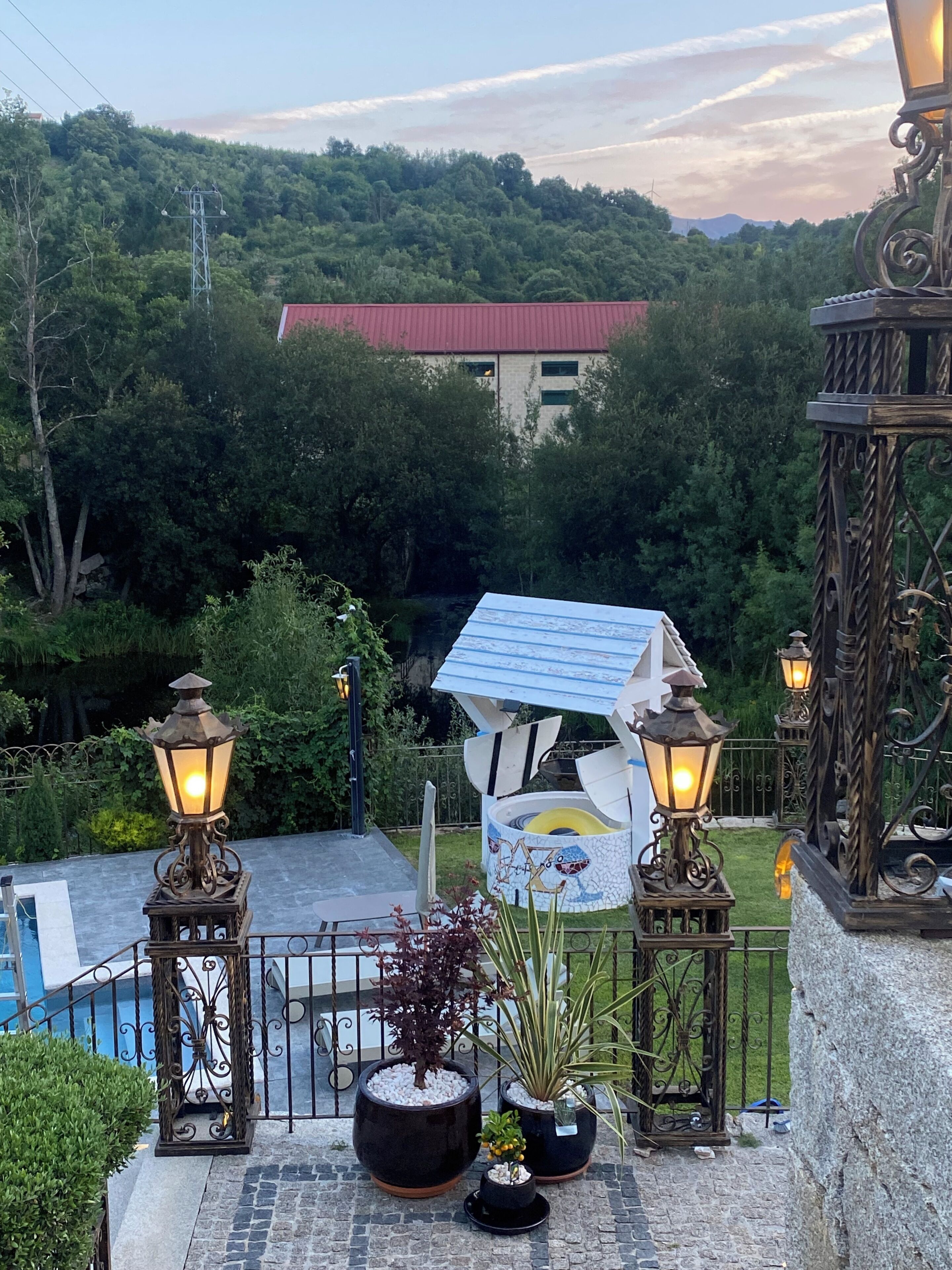 Una terraza en la azotea con vistas a una colina boscosa, un edificio con techo rojo y un farol decorativo.