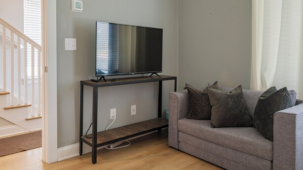 Ein Wohnzimmer mit einem Flachbildfernseher auf einem schwarzen TV-Schrank, einem grauen Sofa mit Kissen und einem Holzboden.