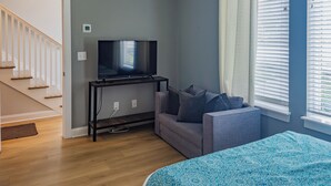 Ein Zimmer mit Fernseher auf einem Schrank, einem grauen Sofa, einer blauen, gemusterten Bettdecke und einer Treppe.
