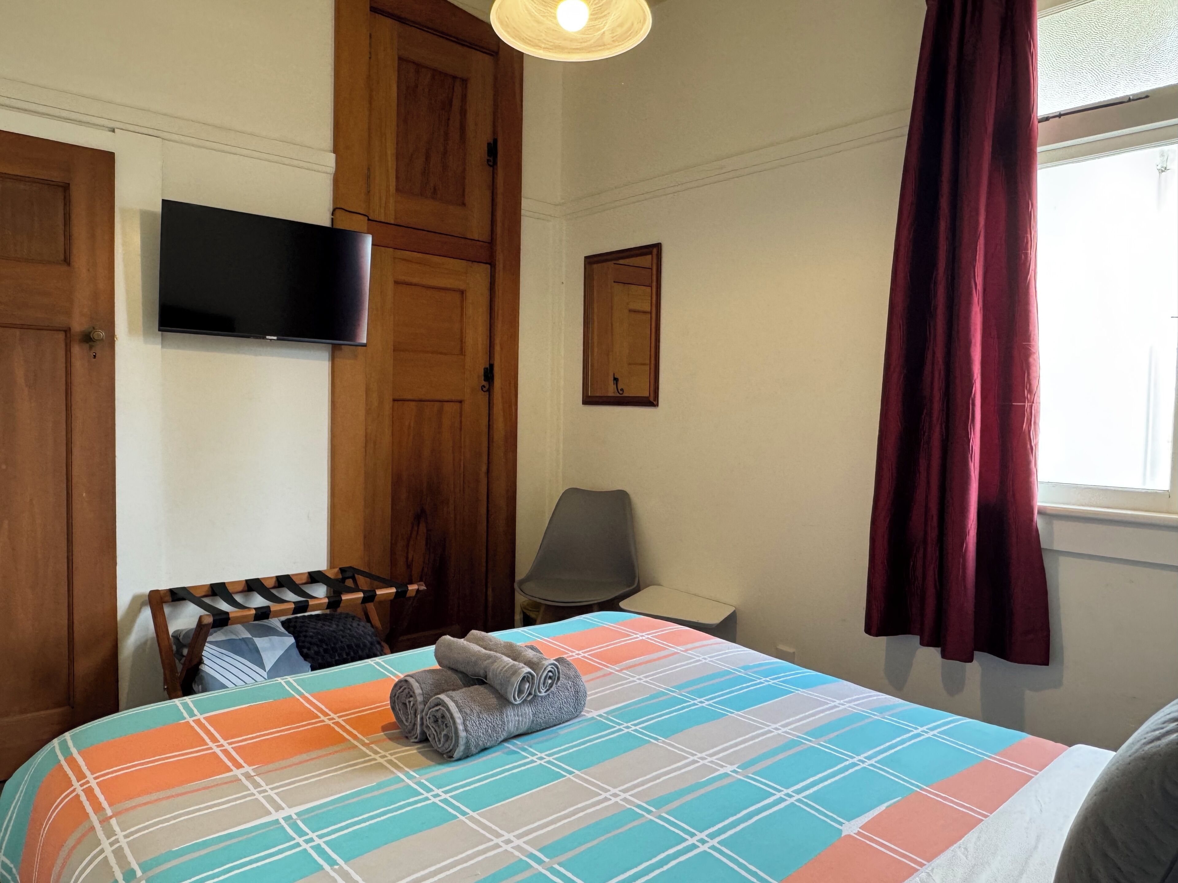 A bedroom with a bed, a television mounted on the wall, a wooden wardrobe, and a window with curtains.