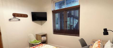 Ein Schlafzimmer mit einem Bett, einem Sessel, einem Fernseher, einem Fenster mit Jalousien und einem kleinen Tisch.