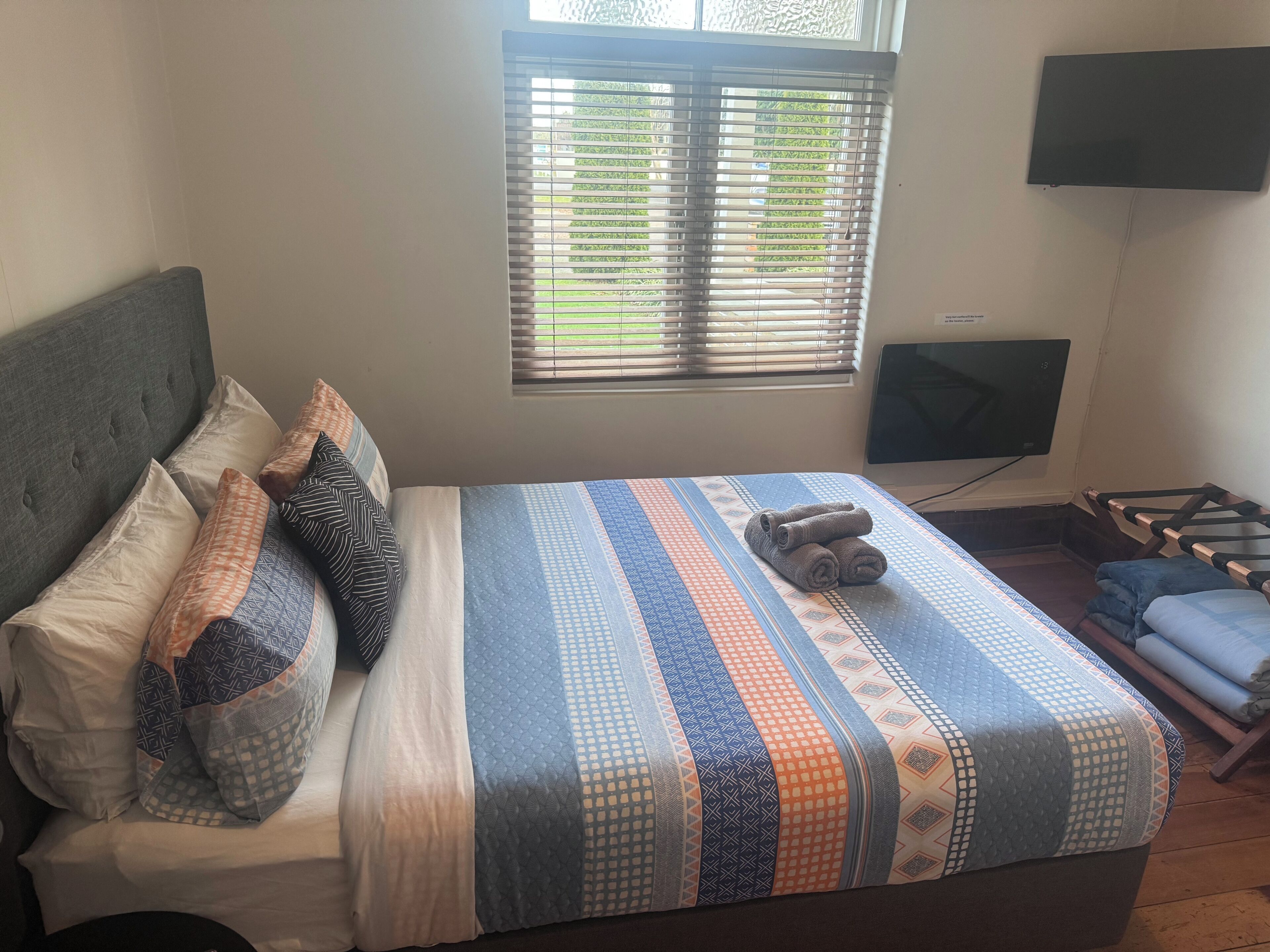 A neatly made bed with a patterned bedspread, a grey upholstered headboard, and a window with blinds.