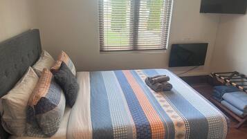 A neatly made bed with a patterned bedspread, a grey upholstered headboard, and a window with blinds.